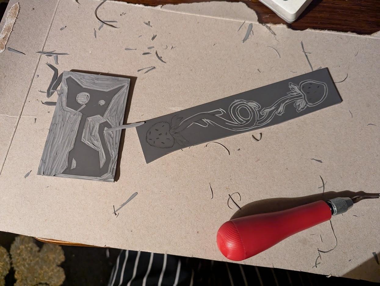 linocut stencils in the process of being carved. one is two strawberries on a vine. the second stencil is a cartoonish cat.