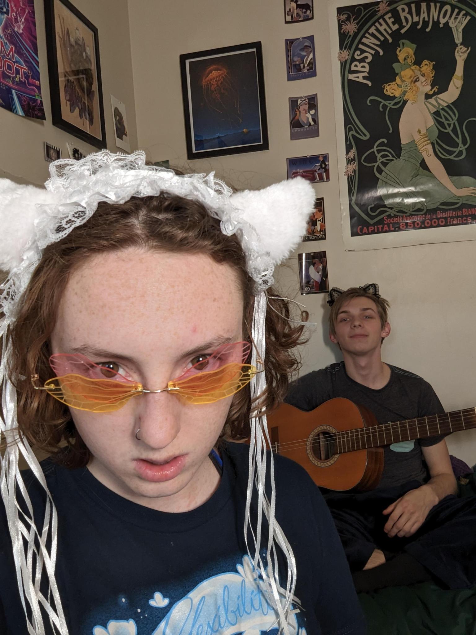 they/them grandson wearing a sheep ear headband and orange sunglasses.  They are looking intensely at the camera. A white guy is in the background holding an acoustic guitar.