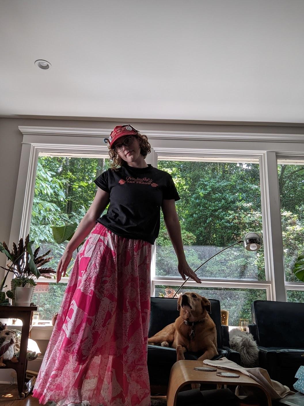 they/them grandson stands in front of a window, wearing a skirt. a dog sits behind them on a chair.